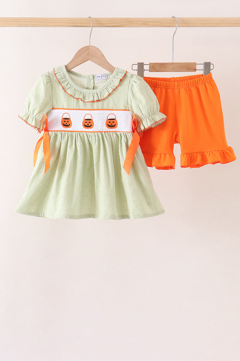 A toddler girl's two-piece Halloween outfit hanging on wooden hangers against a light beige wall. The set includes a light sage green short-sleeve dress with orange ruffle trim, a white chest band with three embroidered orange jack-o'-lantern faces, and matching bright orange shorts with a ruffled hem.